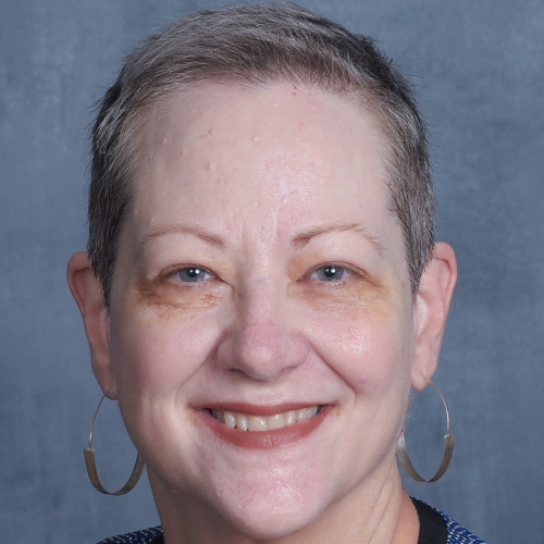 Karen Andes poses in front of a plain grey backdrop wearing a blue textured jacket with black trim and large hoop earrings.